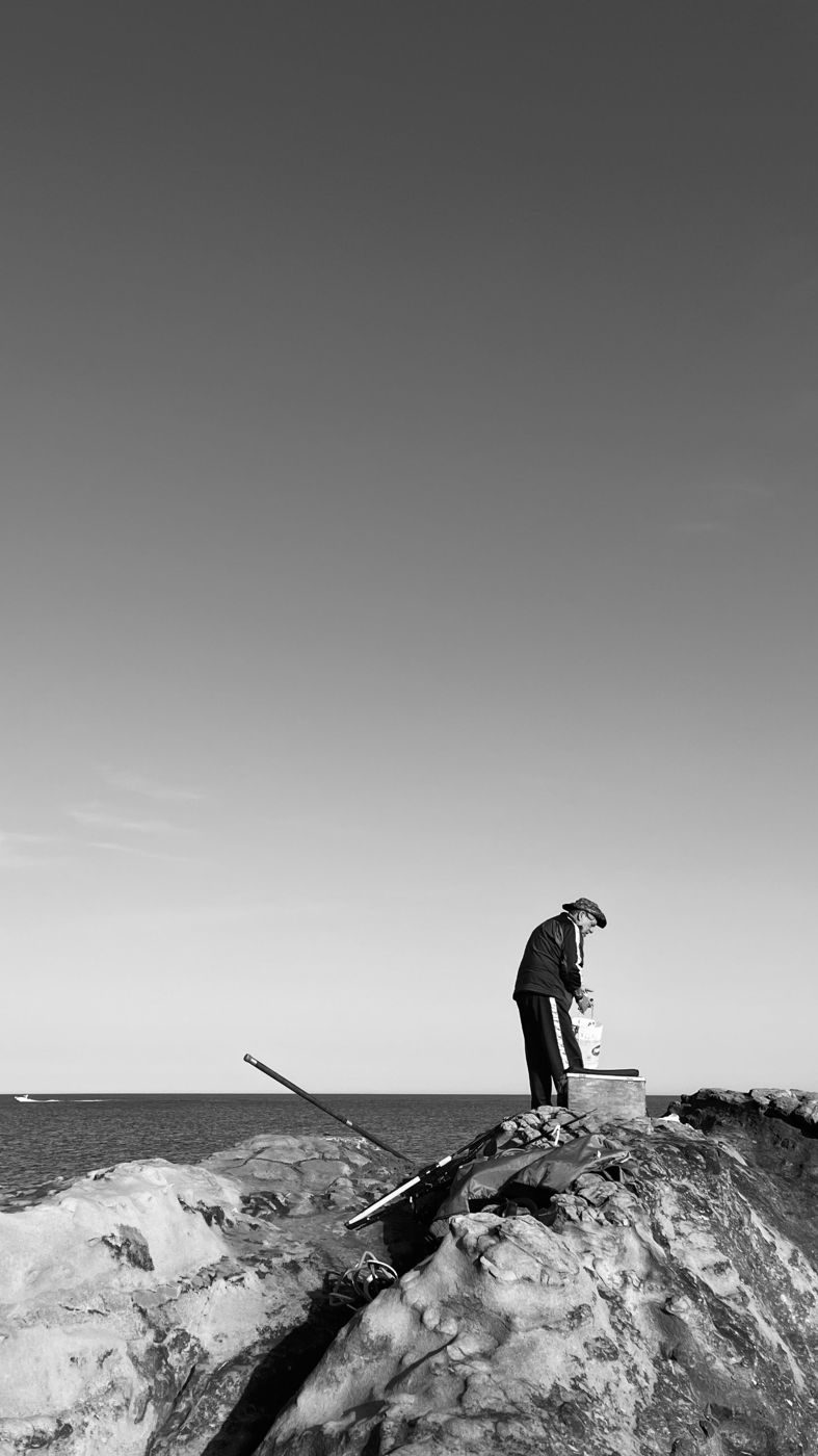 Un pescatore piccolo su uno scoglio, sotto un cielo enorme e vuoto, in bianco e nero
