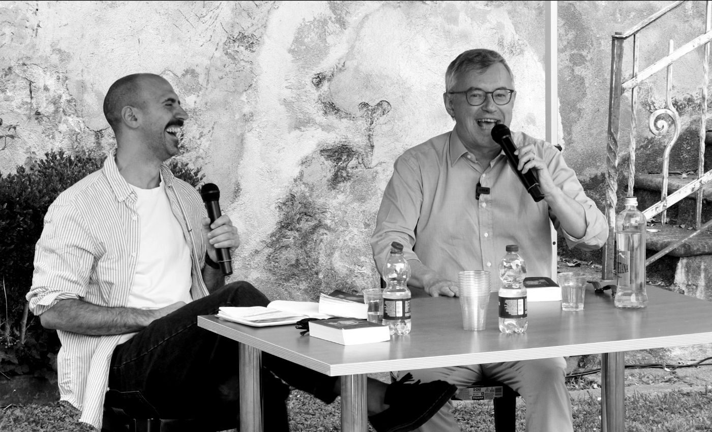 Stefano e Alessandro Barbero ridono durante un incontro pubblico all’aperto, microfoni in mano, libri sul tavolo