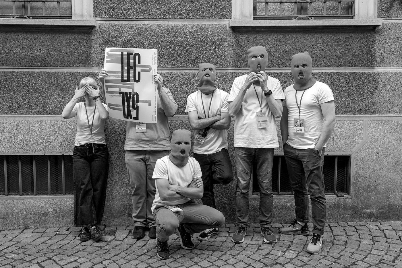 Il collettivo Warde contro un muro, quasi tuttɜ con passamontagna arancioni, uno regge il manifesto della mostra uncovered
